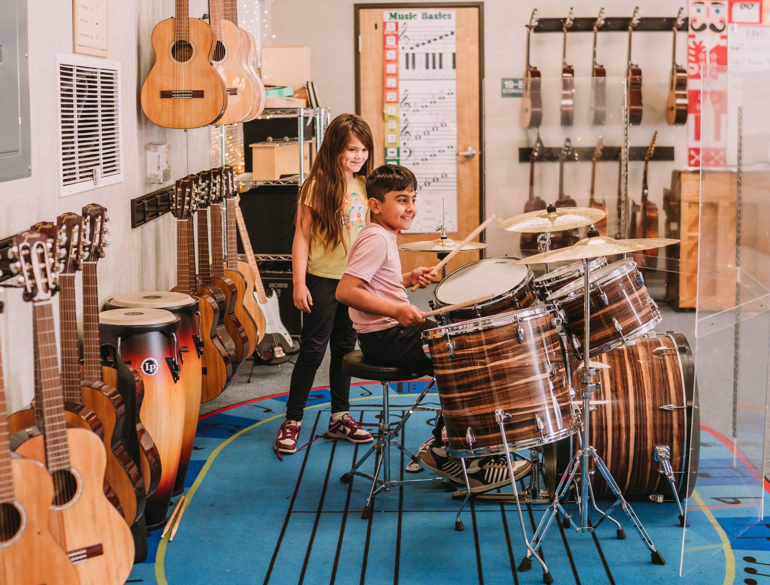 A boy playing the drums while a girls watches. They are surounded by various musical instruments.