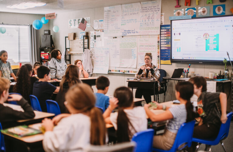 A class of middle school students sitting at their desks activly listenting to the teach who i sin the front of the room teaching a lesson.