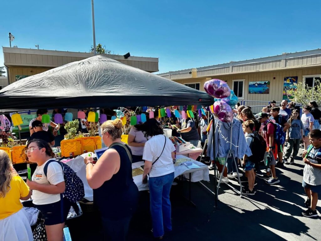 Parent volunteers selling school merchendise and anding out otterpops to students under a popup tent in a sunny day.